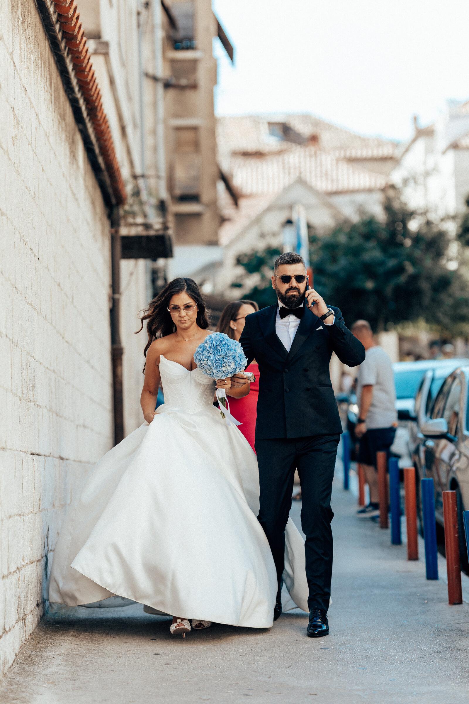 Bride and groom walking along a narrow alley, the bride holding a blue bouquet
