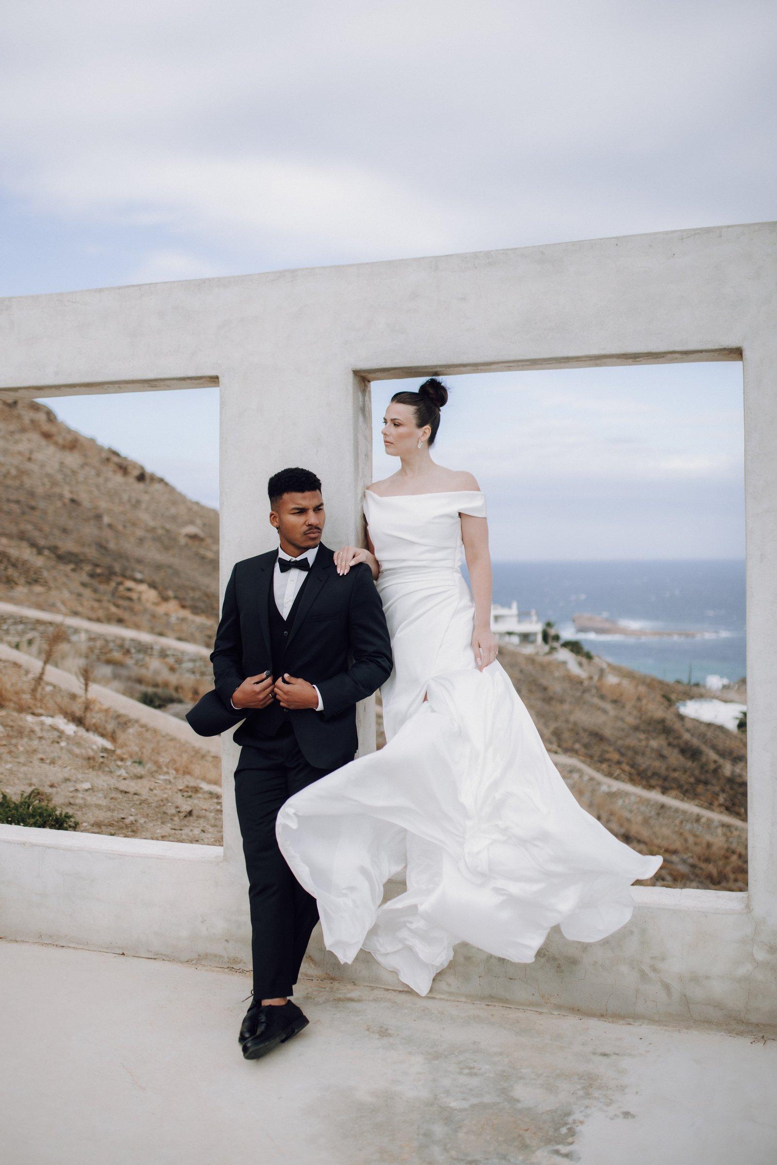 Das Brautpaar vor dem Betonrahmen auf Mykonos, das Brautkleid weht im Wind, dahinter das Meer